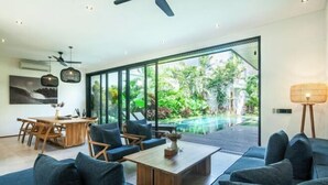 Ein modernes Wohnzimmer mit einem großen Glasfenster zum Poolbereich, einem Essbereich mit Holzmöbeln und einem Deckenventilator.