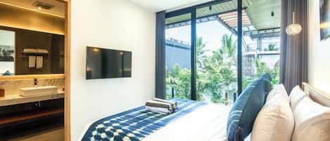 A modern bedroom with a large bed, a built-in TV, a bathroom with a vanity, and a view of greenery through large windows.
