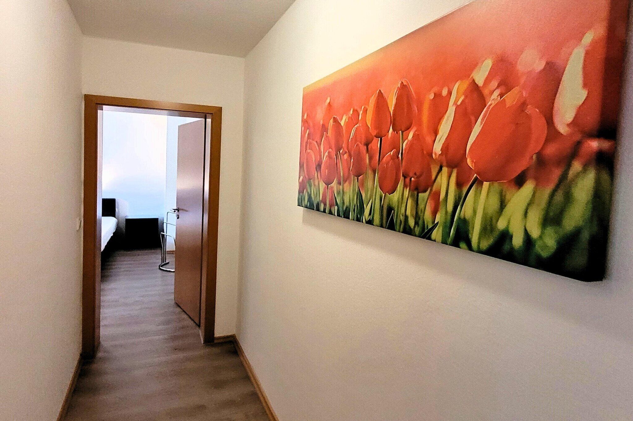 A hallway with a painting of tulips on the wall, a door leading to a room with a bed, and a chair in the distance.