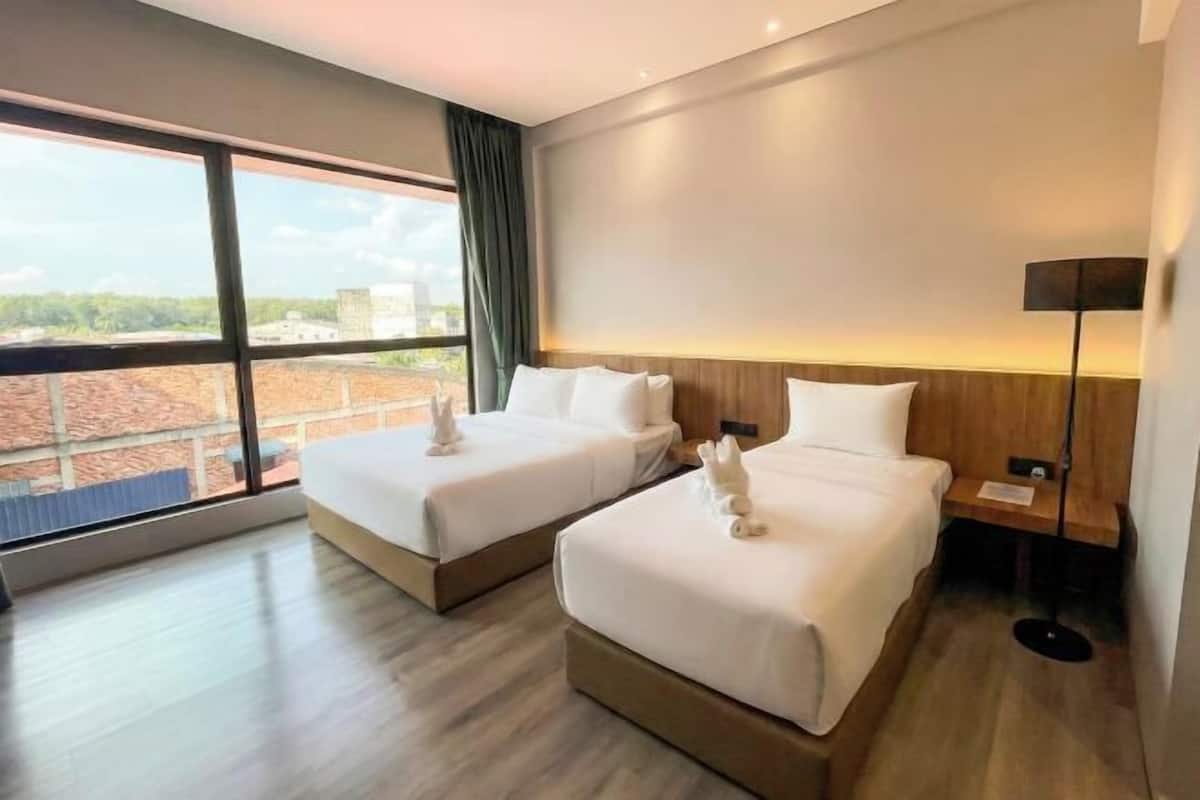 A hotel room with two beds, a large window, a bedside table, and a lamp.
