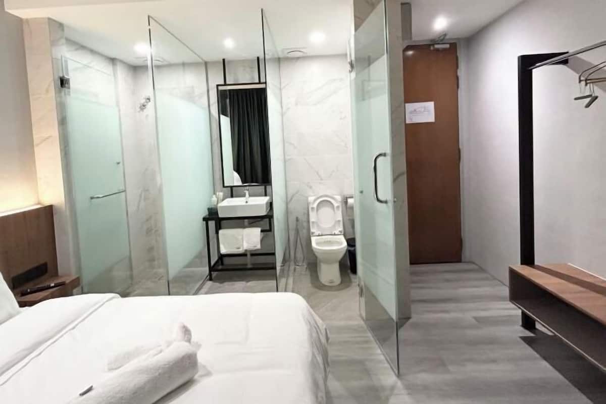 A modern hotel room with a glass-enclosed shower, a separate toilet, a vanity with a mirror, and a bed with a white towel.