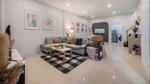 Un salon moderne avec un canapé, des fauteuils, une table basse et un tapis. Des tableaux encadrés ornent le mur, et une lampe se trouve sur une table d’appoint.