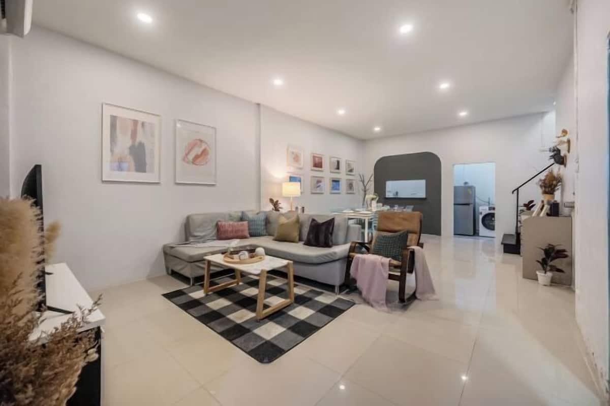 Ein modernes Wohnzimmer mit einer Couch, Sesseln, einem Couchtisch und einem Teppich. An der Wand befinden sich gerahmte Bilder und auf einem Beistelltisch steht eine Lampe.