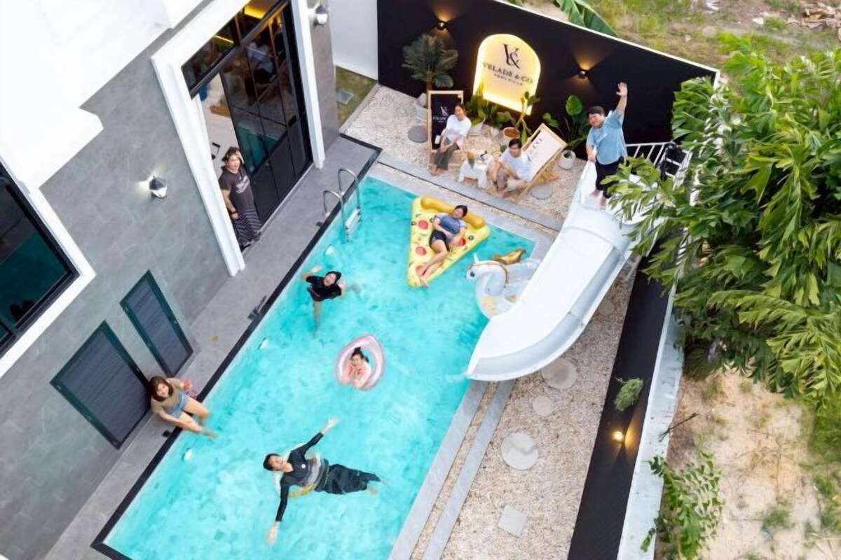 An outdoor swimming pool with people enjoying the water, a slide, and a lounge area with a few individuals.