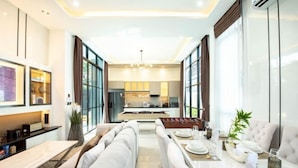 A modern hotel room with a dining area, a sofa, and a kitchen in the background. - Modern 3BR British Pool Villa + 5KM Jomtien Beach (Pattaya)