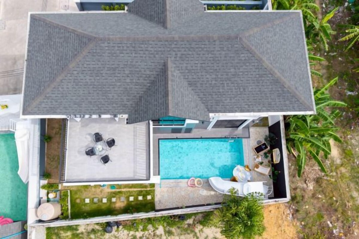 An aerial view of a property with a swimming pool, a covered patio with seating, and a fenced garden area.