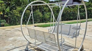 A metal swing set with benches, surrounded by greenery and a building in the background. - Ipoh Homestay Vacation 12 Pax 4 Bedroom for Delicious Food (Ipoh)