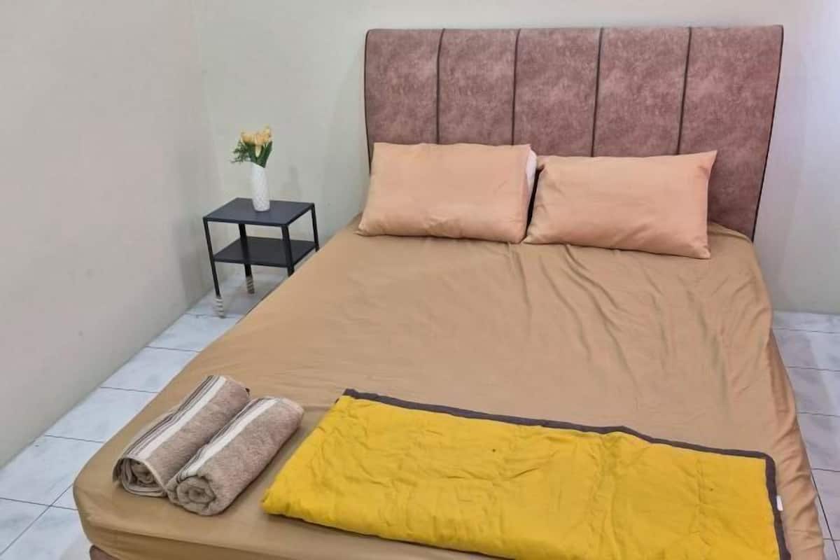 A bed with a headboard, two pillows, a yellow blanket, and two rolled towels on the side.