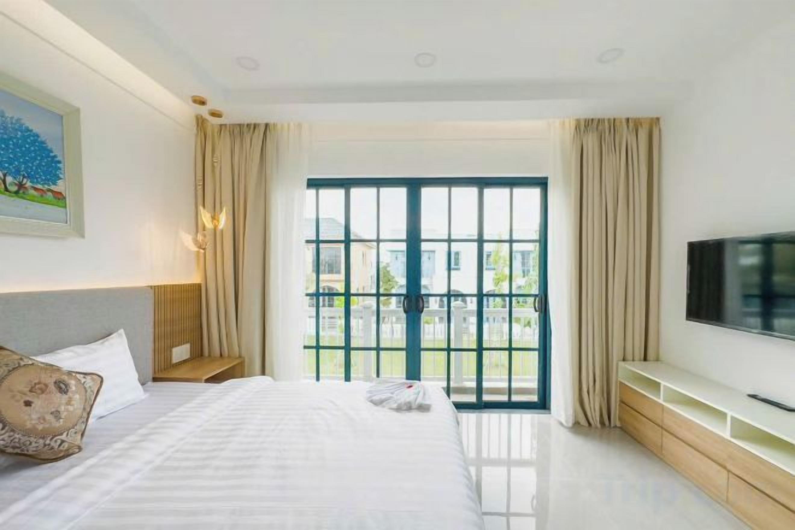 A hotel room with a bed, a TV, a window with curtains, and a painting on the wall.