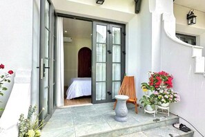 Entrada de hotel con puerta corredera de vidrio, un pequeño espacio de descanso con una silla y una mesa, y plantas en macetas.
