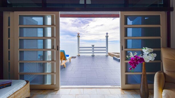 A room with a large open door leading to a balcony with a view of the sea and a white fence.