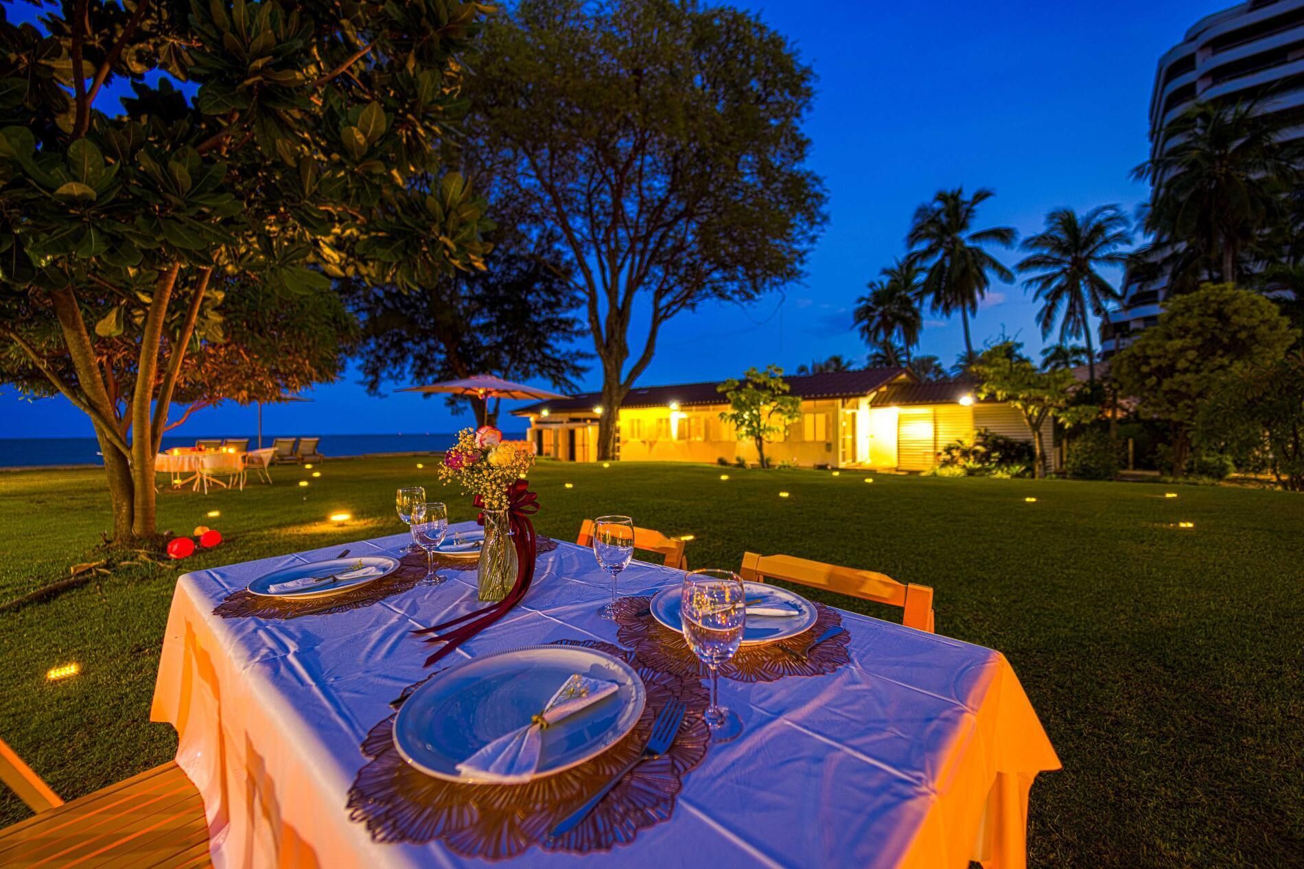 An outdoor dining table set with dinnerware and glasses, surrounded by chairs, with a view of a building and palm trees in the background.