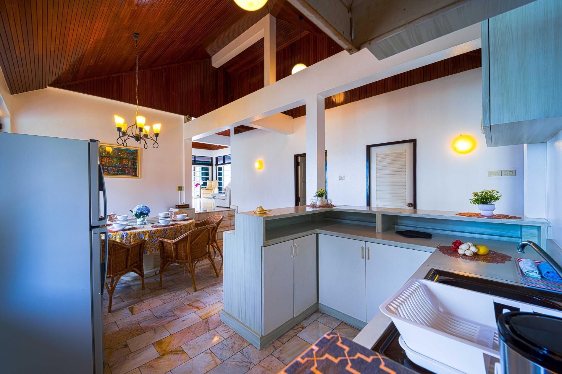 A modern kitchen with a dining area, featuring a refrigerator, dining table with chairs, and a countertop with a sink and fruit bowl.