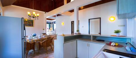 A modern kitchen with a dining area, featuring a refrigerator, dining table with chairs, and a countertop with a sink and fruit bowl.