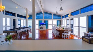 A spacious living area with a pool table, dining table, and chairs. There is a television in the background and large windows providing views of the outdoors.