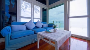 A room with a blue sofa, a white table with a lace tablecloth, and large windows offering a view of the sea.