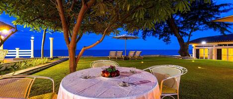 Un área de comedor al aire libre con una mesa para dos, rodeada de sillas, con vista a una playa al atardecer.