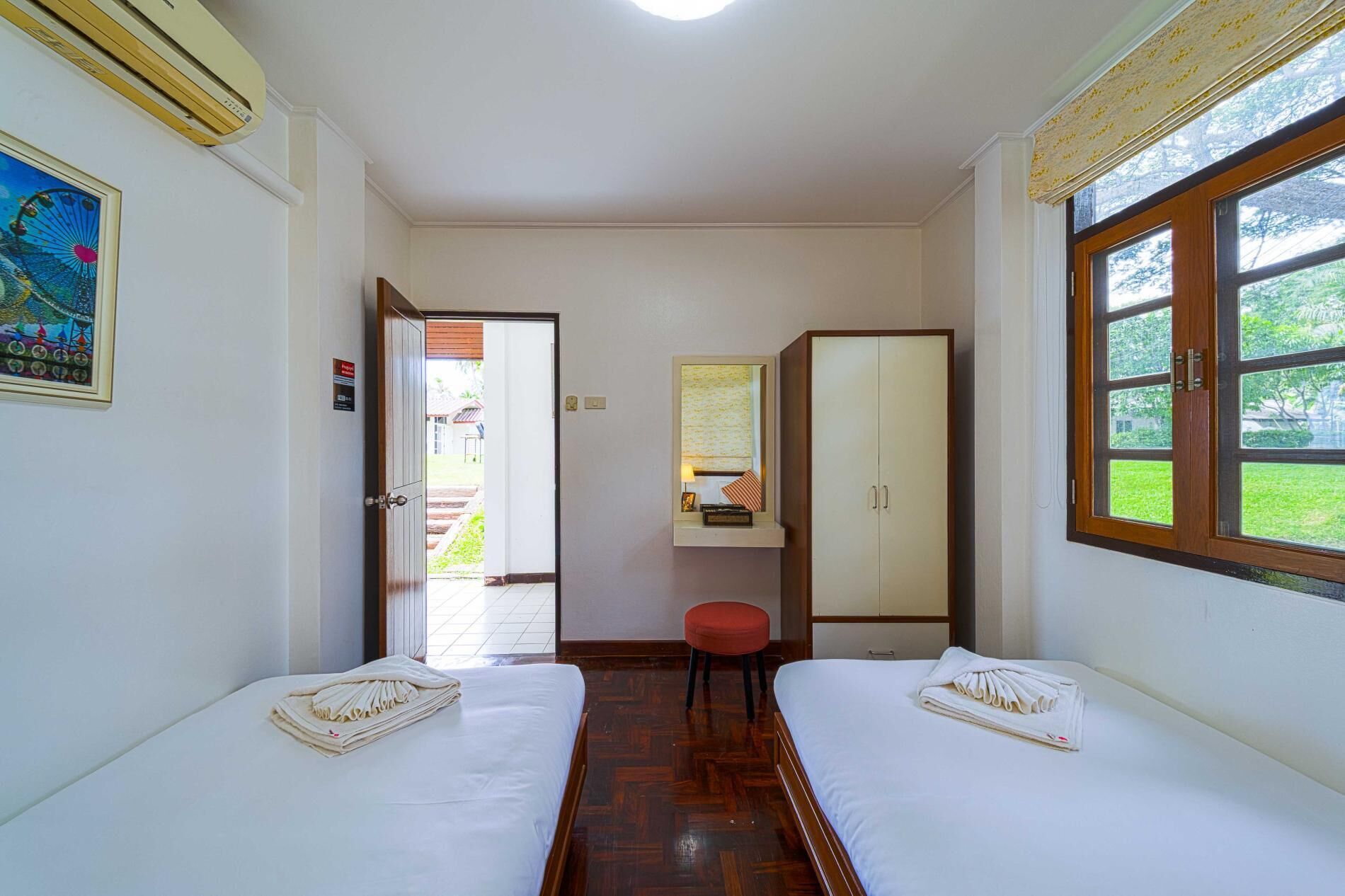 Una camera d'albergo con due letti, un armadio, uno sgabello e una finestra vista verde.