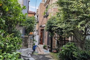 A narrow alleyway with a person tending to plants, surrounded by dense foliage and residential buildings. - MingYue Building (Tokyo)