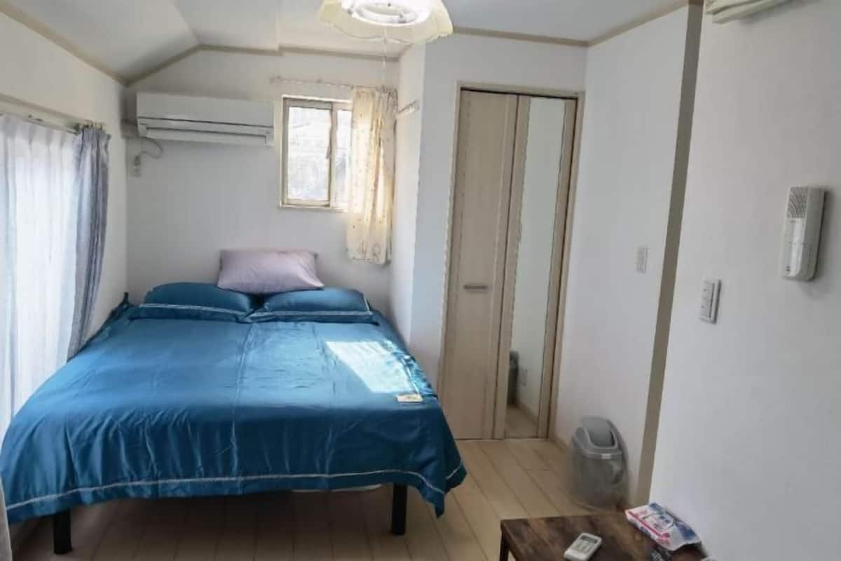 A bedroom with a bed, curtains, an air conditioning unit, a window, a door, a trash bin, and a small table with a remote control.