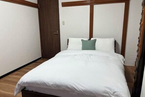 Un lit simple avec une literie blanche, dans une pièce aux boiseries apparentes et dotée d’une porte.