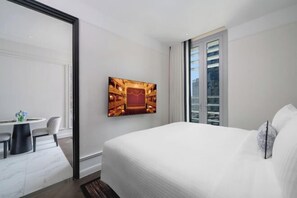 A hotel room with a large bed, a dining table with chairs, a painting on the wall, and a view of the city through the window.