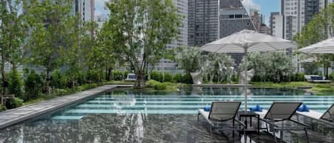 A rooftop pool with lounge chairs and umbrellas, surrounded by greenery and city buildings.