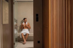 A man sitting in a shower with a towel, a glass door, and wooden paneling. - La Clef Bangkok by The Crest Collection (Bangkok)