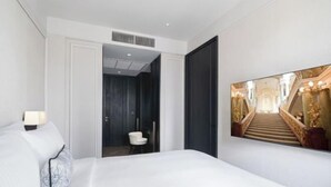 A hotel room with a bed, a nightstand with a lamp, a chair, a door leading to another room, and a large framed picture on the wall. - La Clef Bangkok by The Crest Collection (Bangkok)