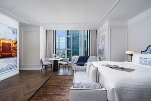 A hotel room with a large bed, a seating area with two chairs, a small table, and a view of the city through large windows.