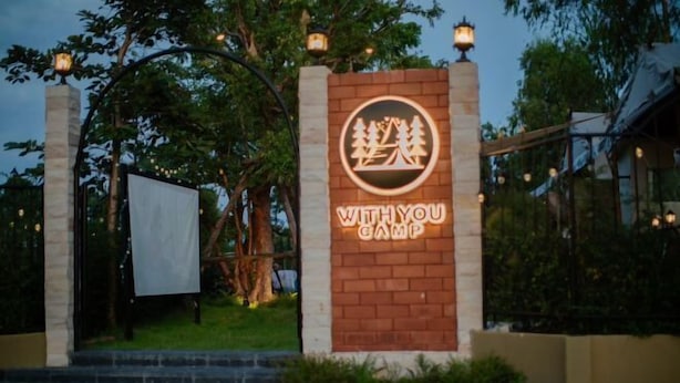 A sign for "With You Camp" with a campfire and trees logo, surrounded by greenery and a brick wall. - With You Camp (Sirindhorn)
