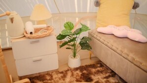 A modern bedroom with a bed, a bedside table with a lamp, a plant, and a fluffy rug. - With You Camp (Sirindhorn)