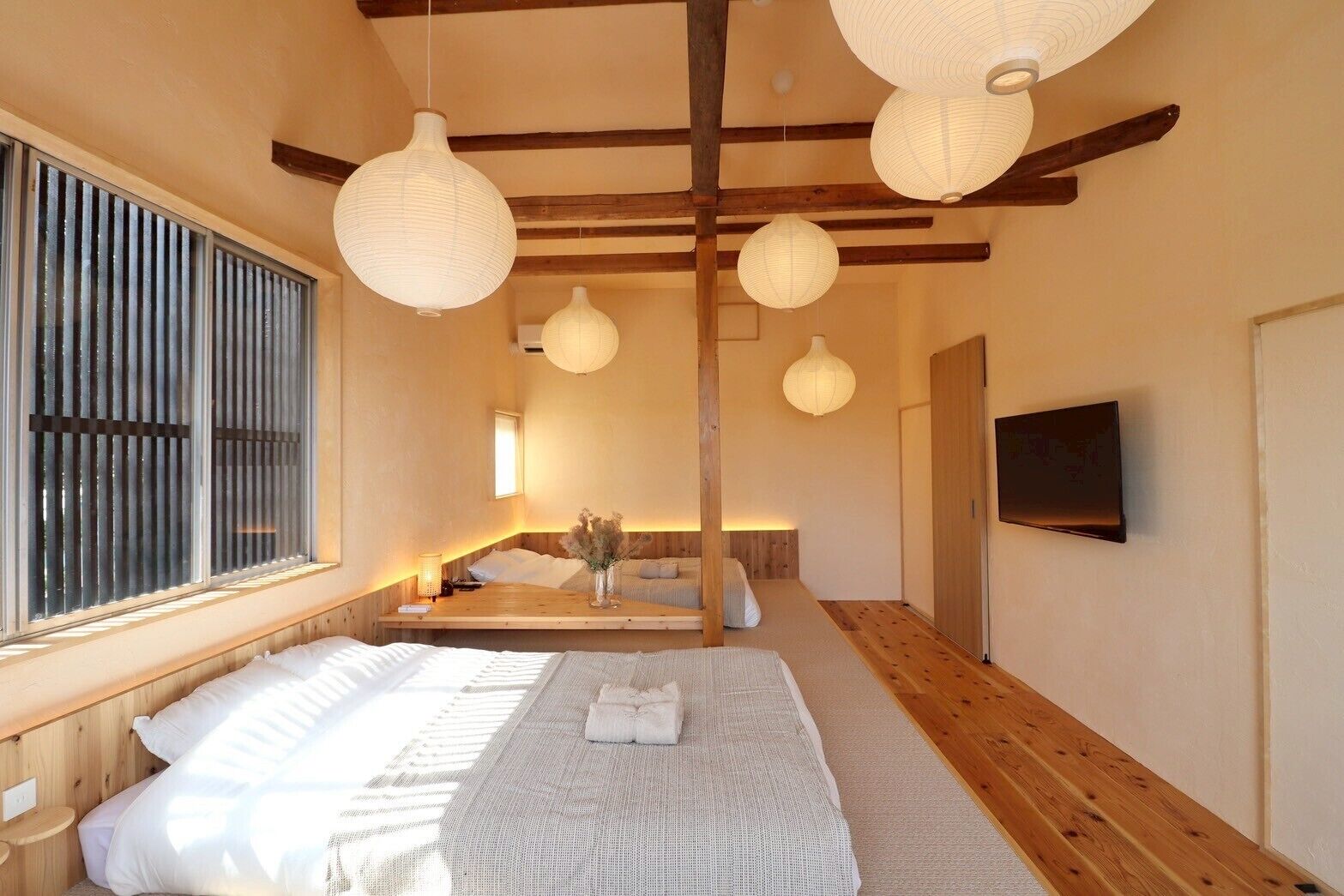 A room with a bed, a bedside table, a window with blinds, and a television mounted on the wall.