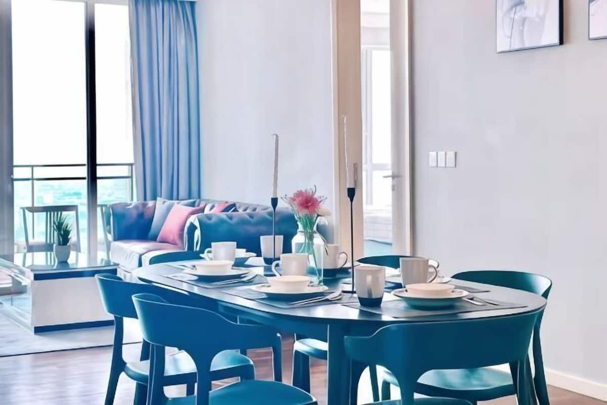 A dining area with a table set for four, a sofa in the background, and a view of the outdoors.