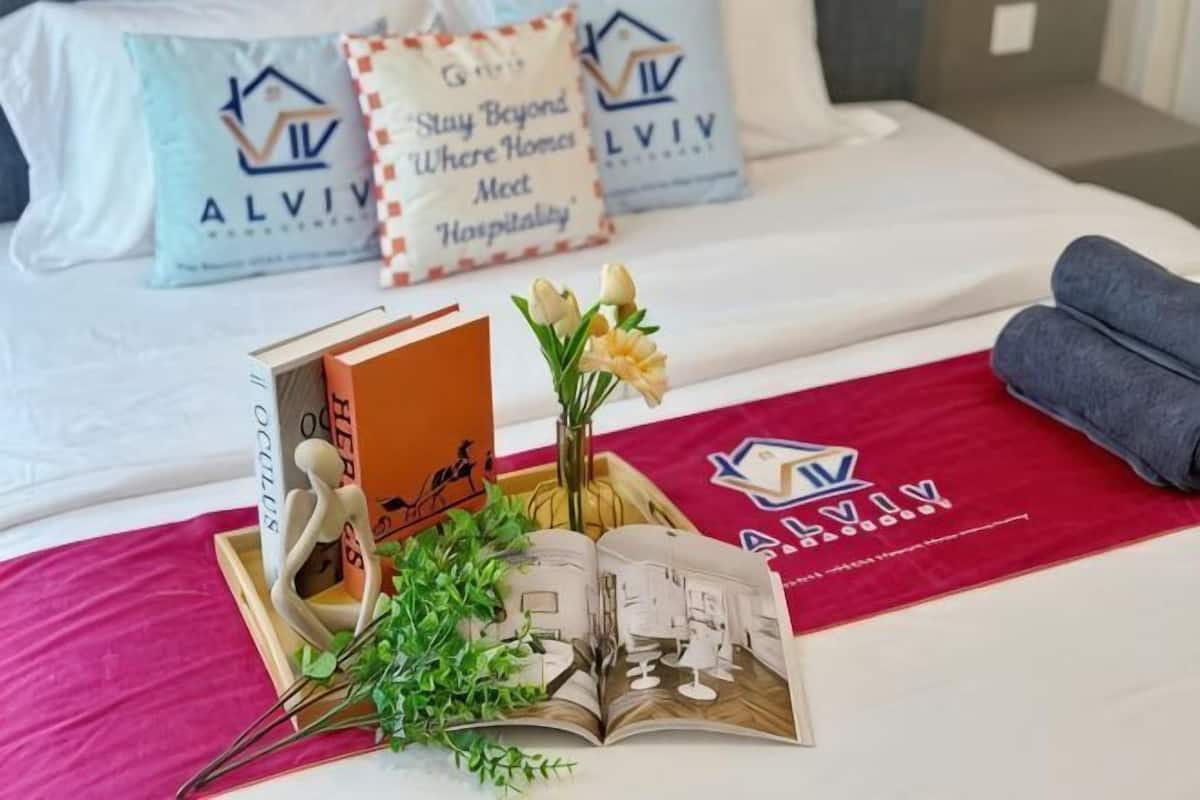 A neatly made bed with pillows, a book, a vase with flowers, and a red blanket with a logo.
