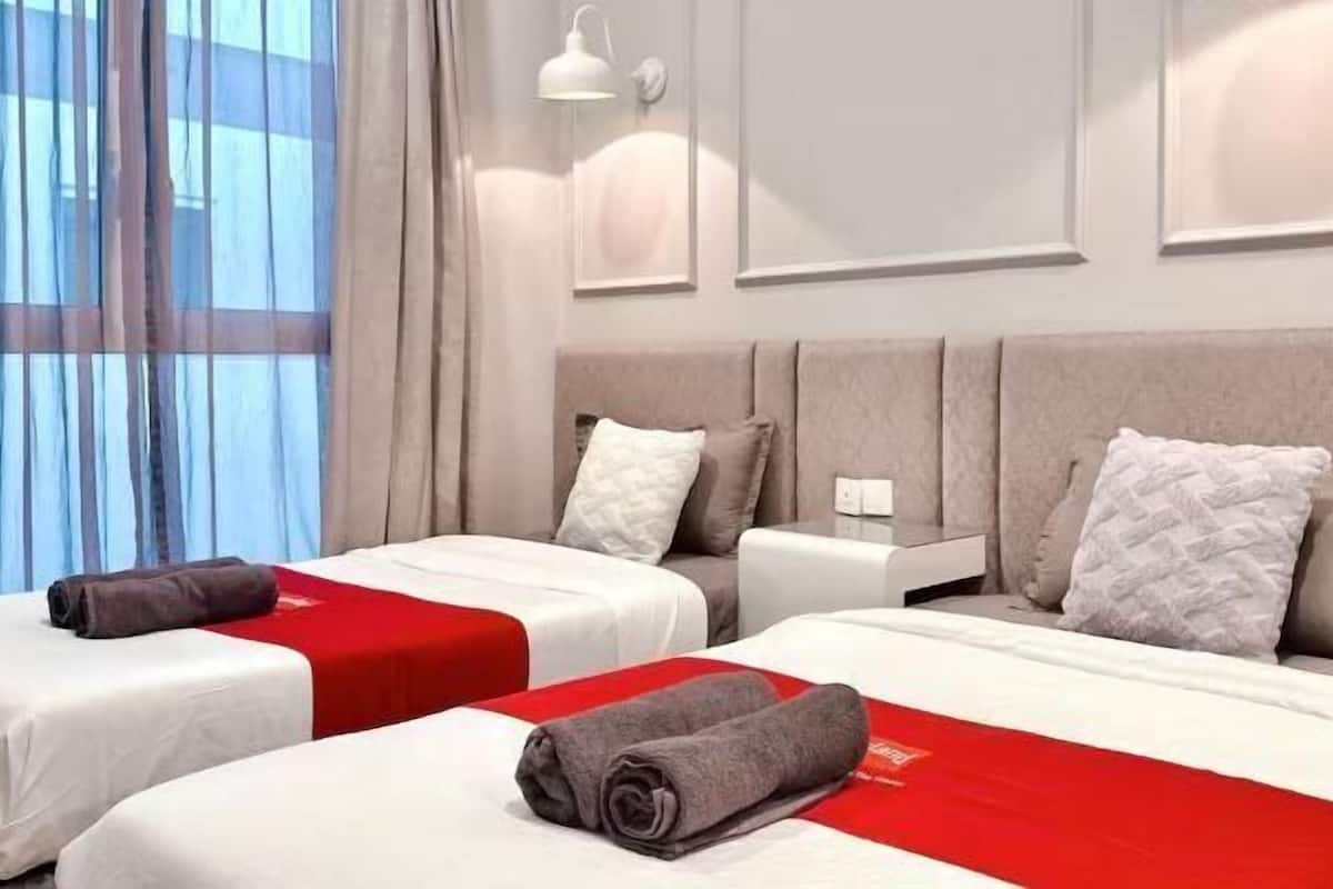 A hotel room with two beds, a large window with curtains, a wall-mounted lamp, and a headboard with pillows.