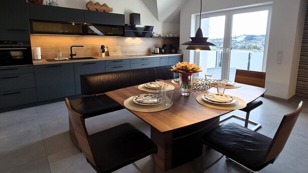 A modern kitchen with dark cabinetry, a wooden dining table set for four, and a large window offering a view of the city. - Duplex in Winterberg near Market Square (Winterberg)