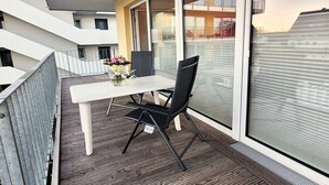 Un balcon avec une table blanche et des chaises noires, un vase de fleurs et une vue sur un immeuble doté de balcons.