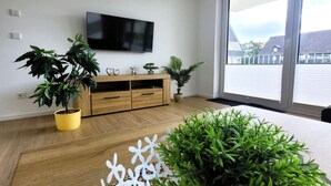 Un salon moderne avec un plancher en bois, un téléviseur à écran plat fixé au mur, un meuble en bois et des plantes en pot.