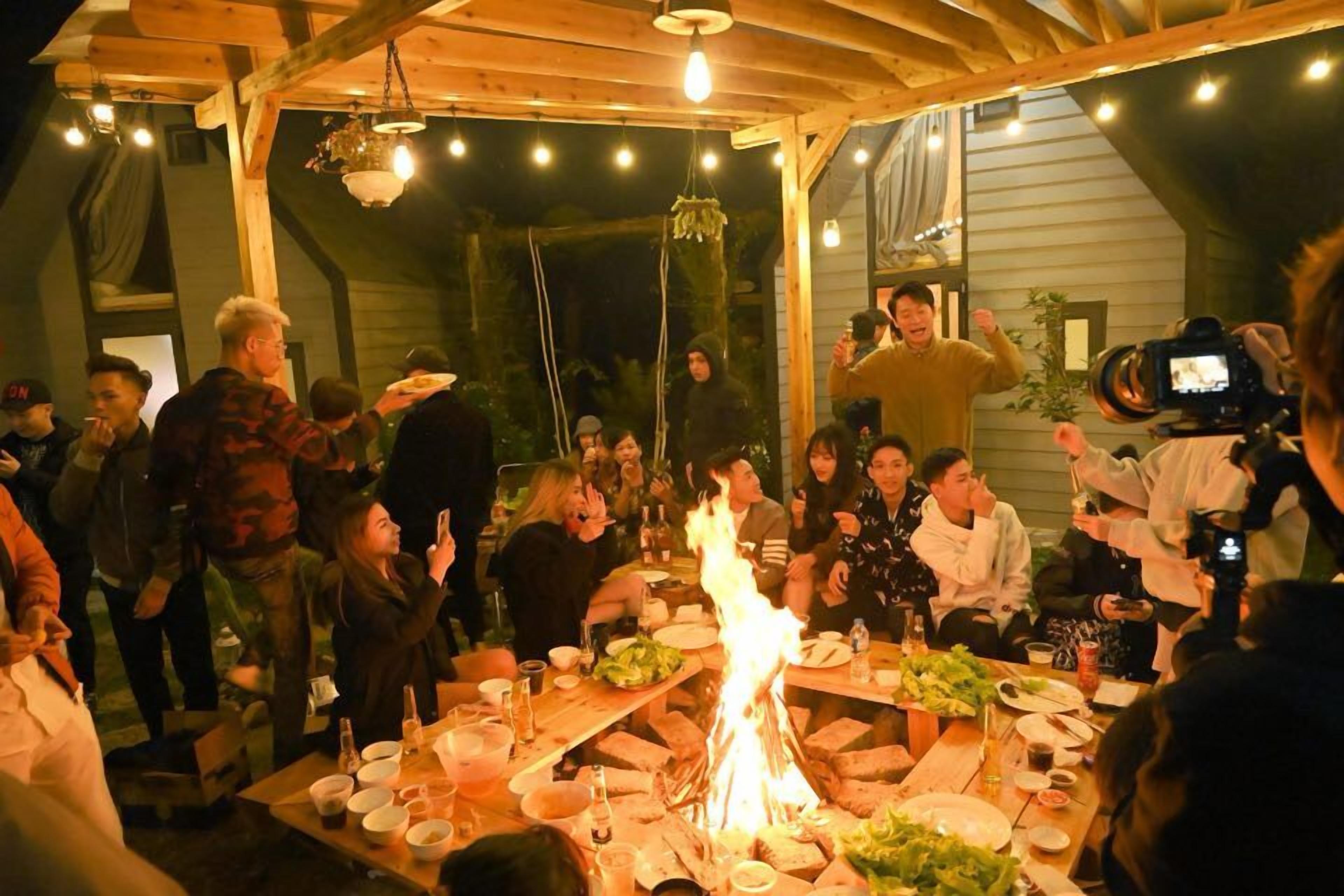 A group of people gathered around a table with a fire, dining and socializing in a rustic outdoor setting.