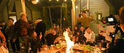 A group of people gathered around a table with a fire, dining and socializing in a rustic outdoor setting.