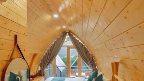 A cozy attic bedroom with a wooden sloped ceiling, a large window, a bed with pillows, a mirror, and a hanging coat rack. - The Chill Garden Sapa (Ta Van)