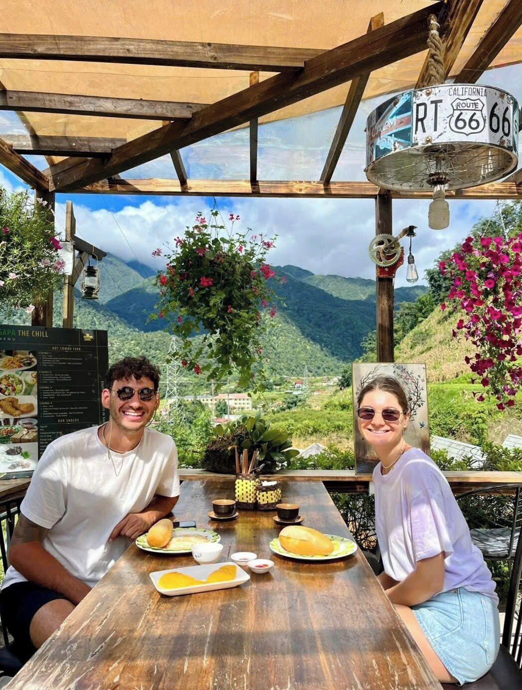 Two people seated at a wooden table with a menu, surrounded by a scenic mountain view and a sign indicating a route number.