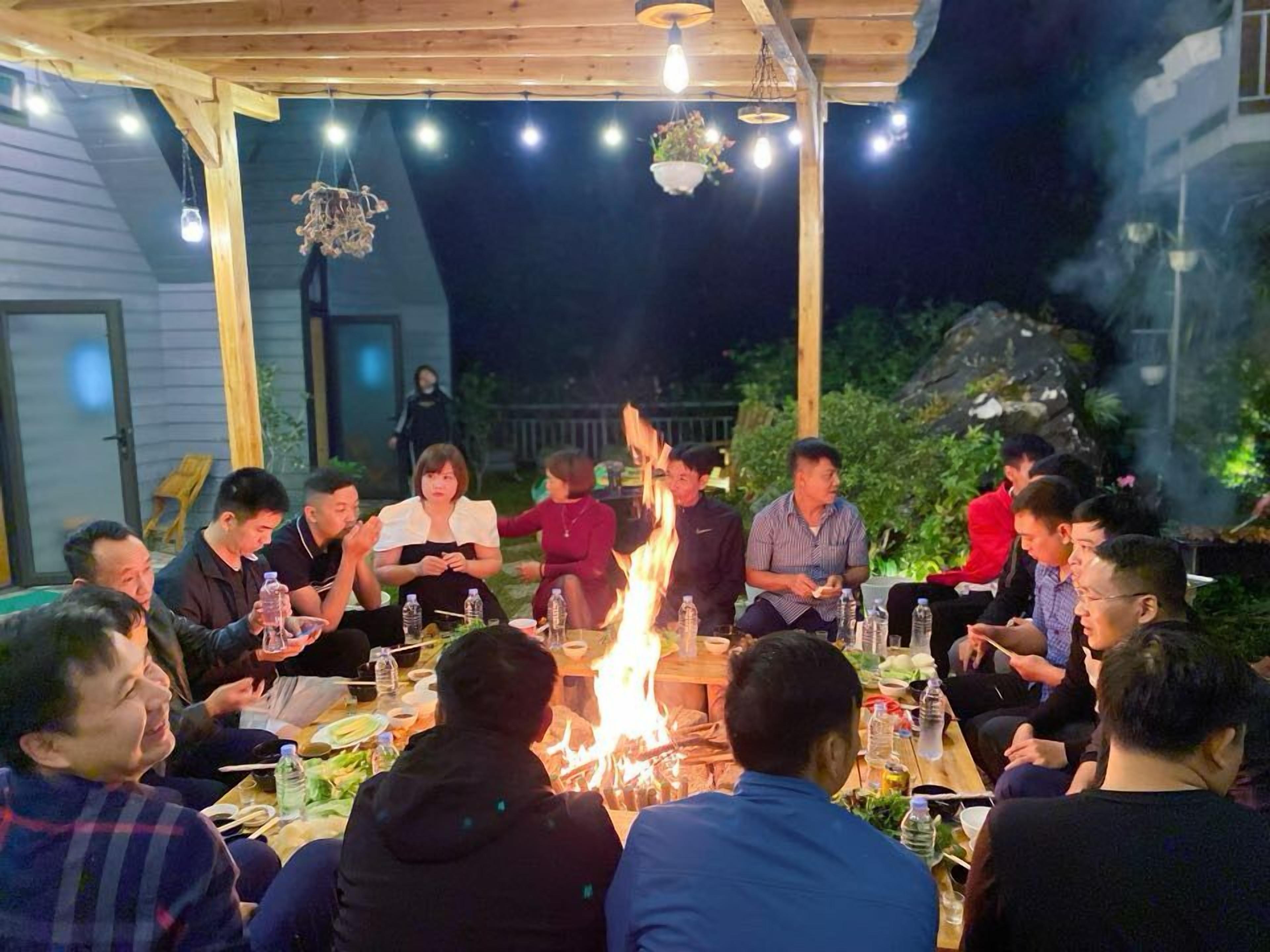 A group of people gathered around a fire pit outdoors at night, with tables set for a meal.