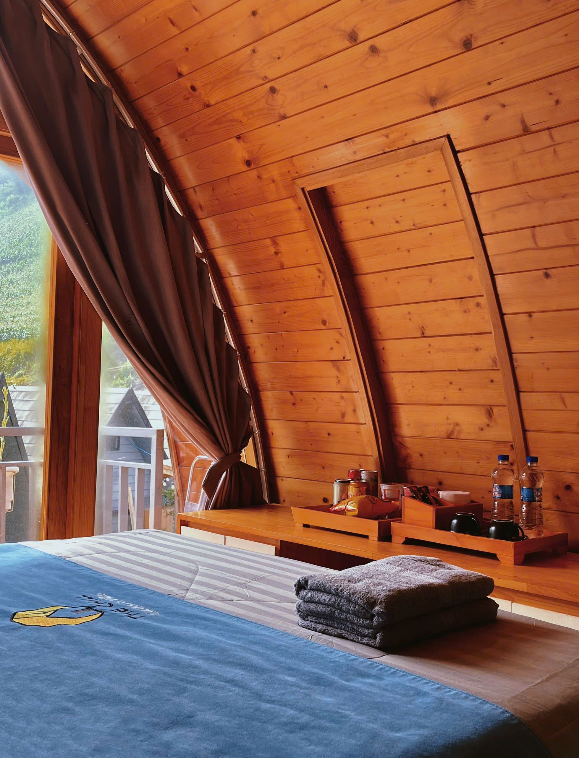 Une chambre confortable située dans les combles, avec un plafond en pente, un lit recouvert d’une couverture bleue, un banc en bois où sont posés des articles de toilette, et une vue sur l’extérieur.