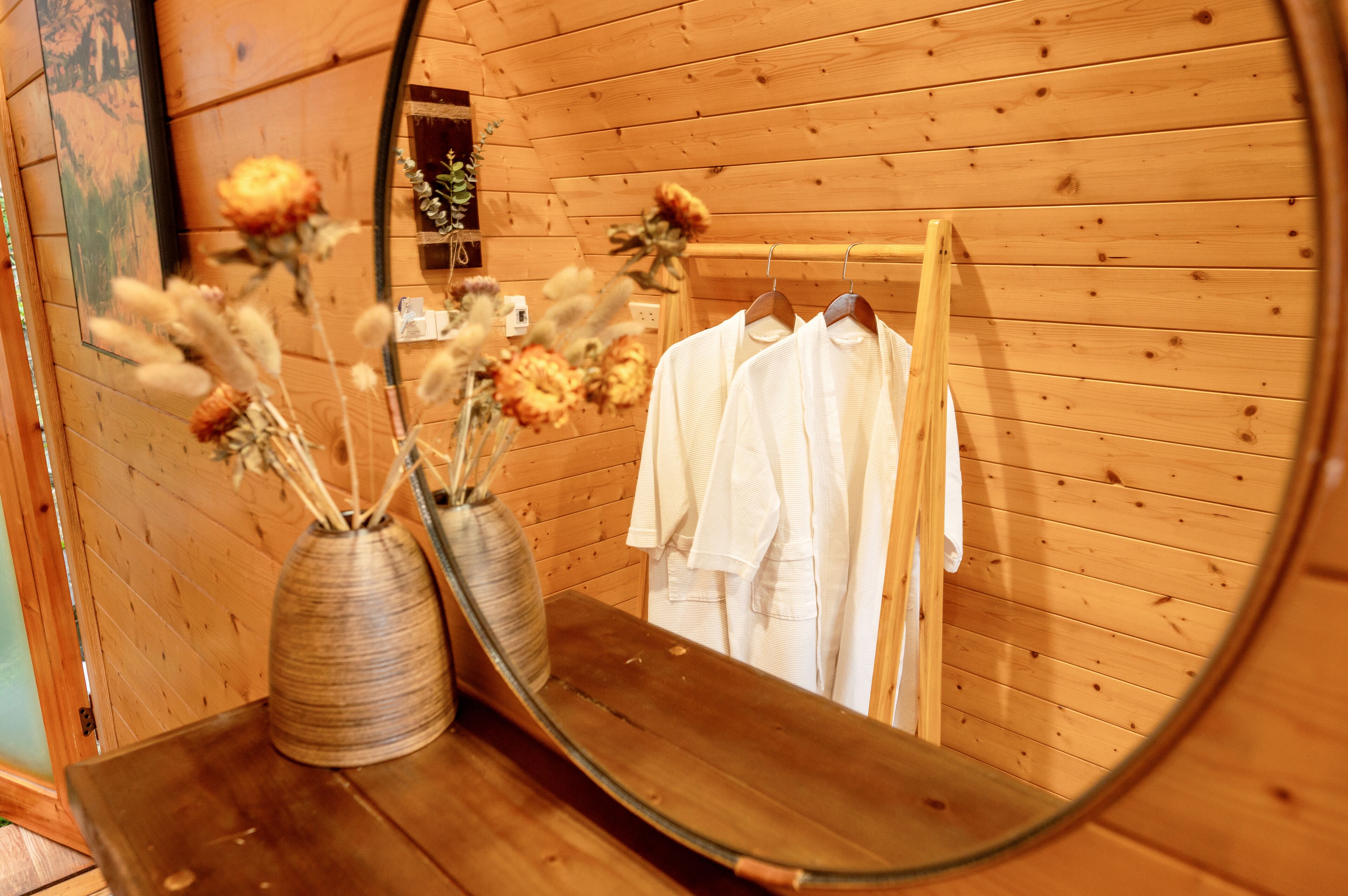 A wooden interior with a mirror reflecting a white shirt and a vase with dried flowers.