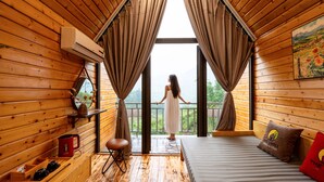 A wooden cabin room with a bed, a bench, a window with curtains, and a view of the outdoors. - The Chill Garden Sapa (Ta Van)