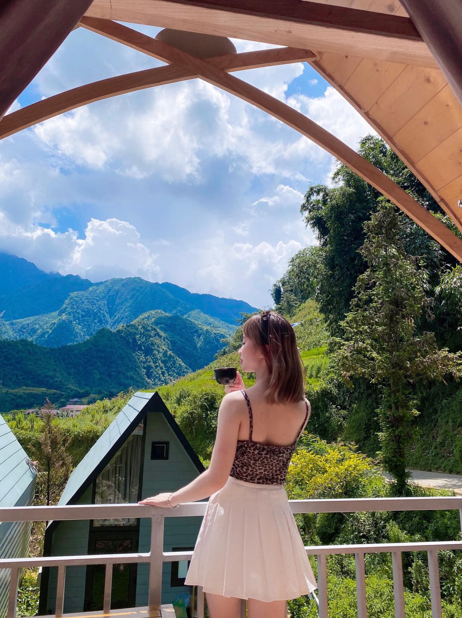 A woman standing on a balcony with a view of green hills and a house in the background.