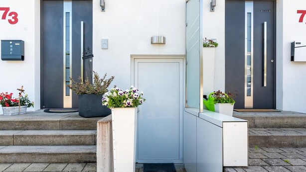 A building entrance with two doors marked with the number 73, a small step leading up to the entrance, and potted plants on either side.