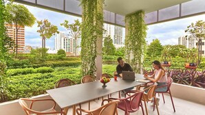 Una mesa con sillas, una computadora portátil y un frutero en una habitación con ventanas amplias y plantas.